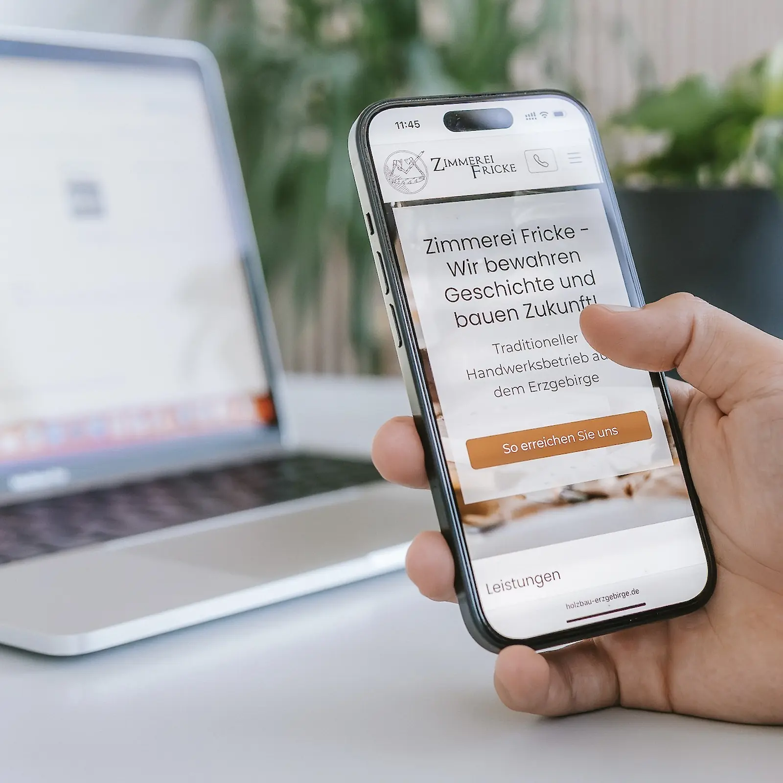Eine Hand hält ein Smartphone, auf dem die Website der Zimmerei Fricke angezeigt wird, mit dem Slogan „Wir bewahren Geschichte und bauen Zukunft“. Im Hintergrund sind ein unscharfer Laptop und eine grüne Pflanze zu sehen.